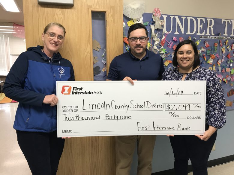 Check presented by Trevor Brittsan (center) of First Interstate Bank to Shelley Moore (left), Principal of Sam Case, and Kristin Becker (right), Principal of Yaquina View Elementary. Standing in front of a Sam Case classroom in need of a Lockdown Shade.